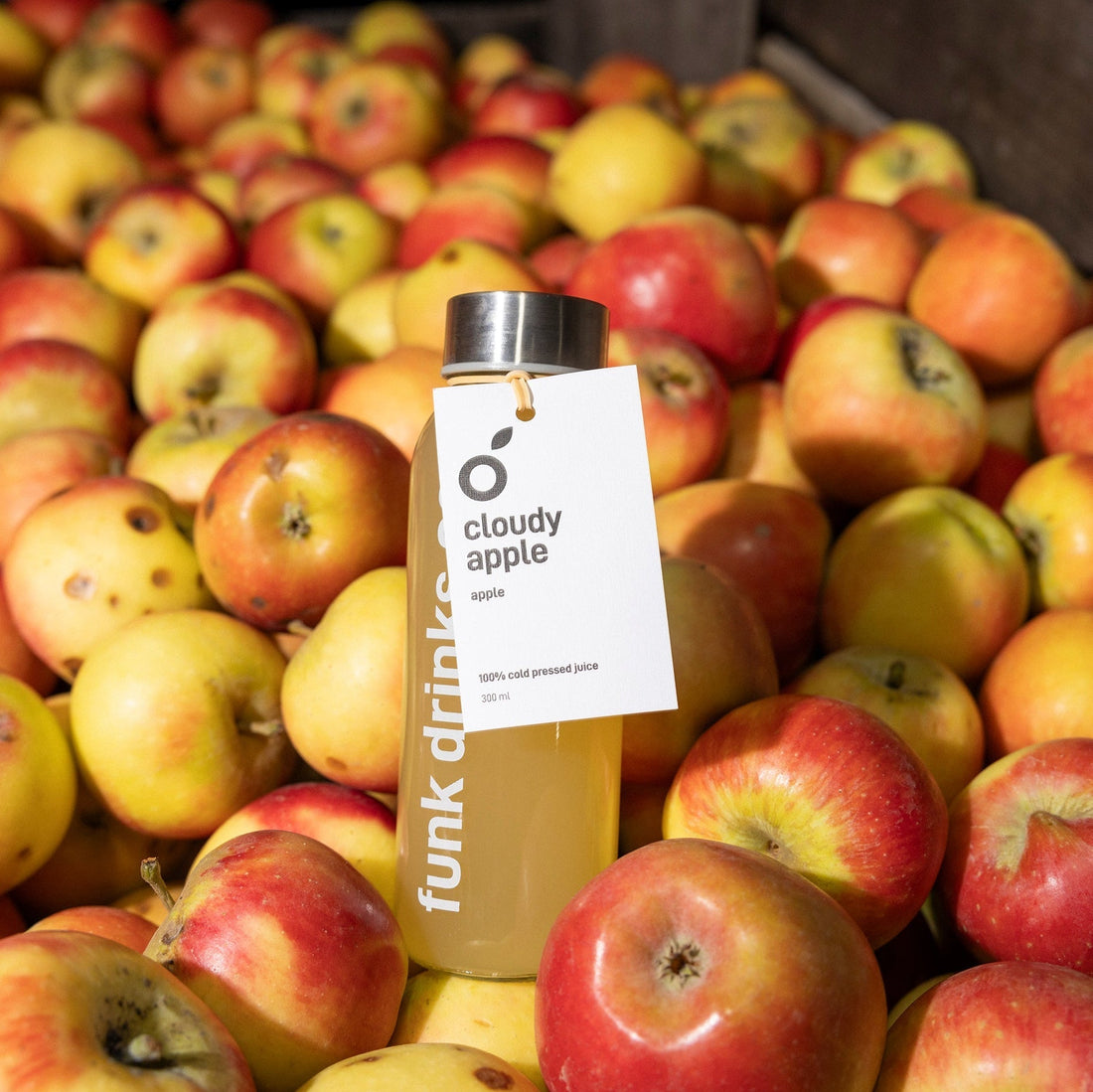 A bottle of Funk Drinks Co Cloudy Apple cold pressed juice standing in a crate of freshly picked red and yellow apples