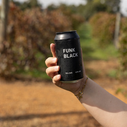 A hand holding a can of Funk Black dark ale in front of the vines